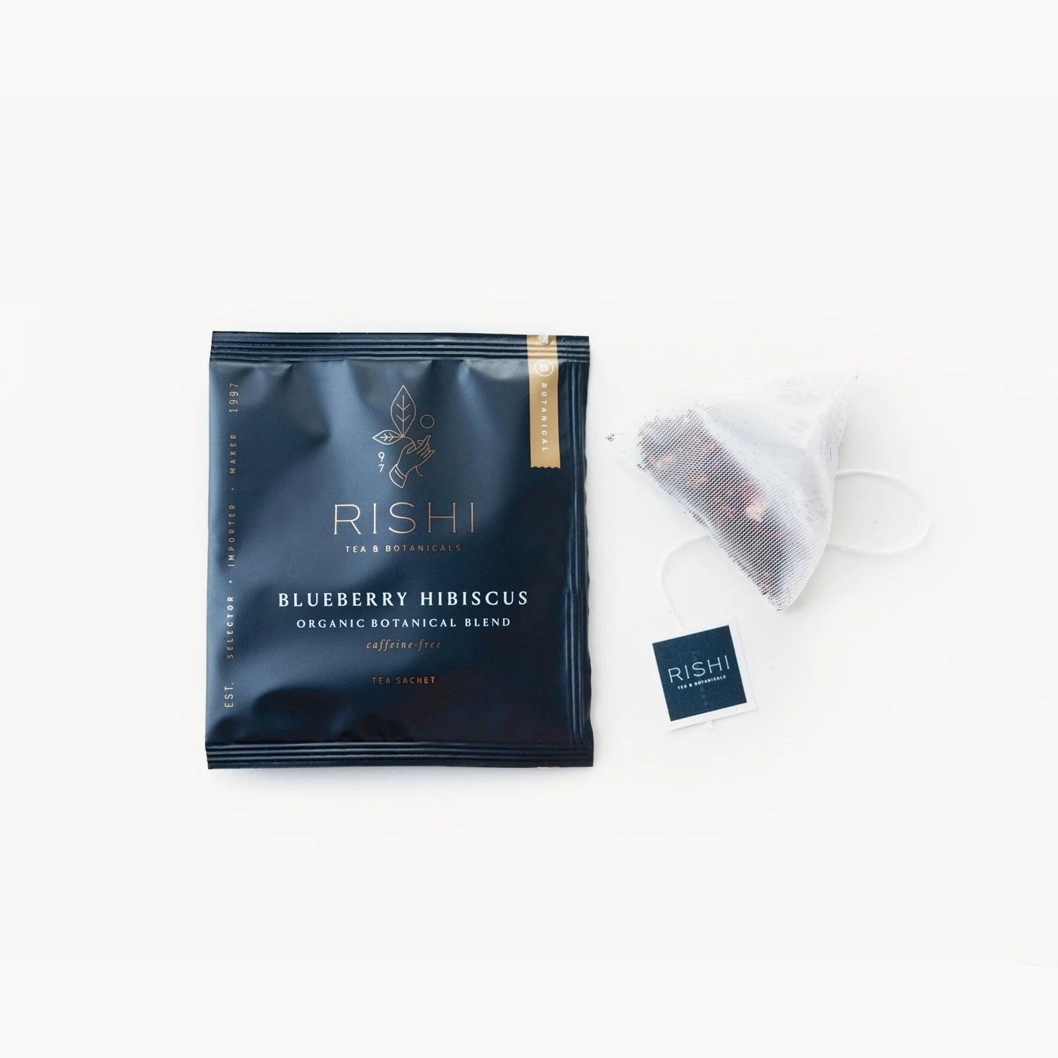 A tea sachet package with the label 'RISHI' and 'BLUEBERRY HIBISCUS' text, alongside an individual sachet partially unfolded to show the tea inside.