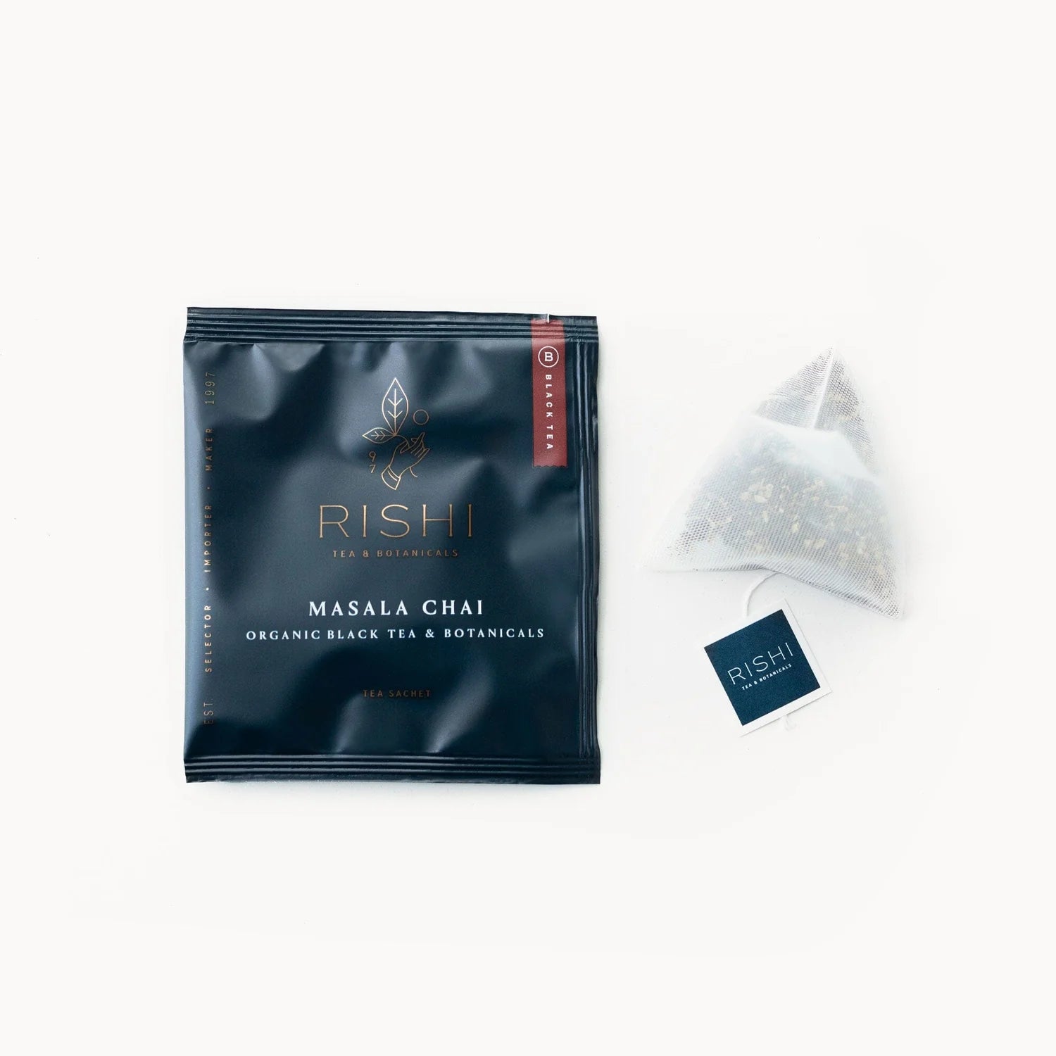 A black tea sachet packet with the brand name RISHI and the product name Masala Chai visible, alongside a clear plastic sachet showing the tea leaves inside.